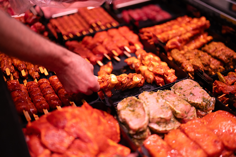 Ausgelegtes rohes gewürztes Fleisch und Fleischspieße in einem Metzgerei-Display mit einer Hand, die einen Spieß aufnimmt.