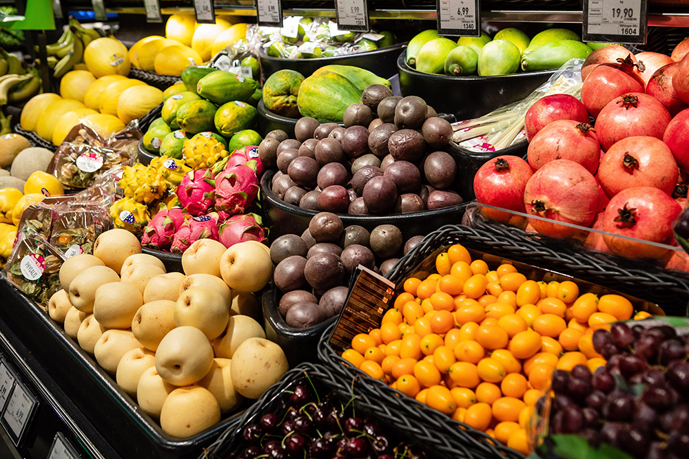 Verschiedene bunte Früchte in Körben auf einem Marktstand, darunter Granatäpfel, gelbe Kirschen, Drachenfrüchte, grüne Papayas und gelbe Melonen.