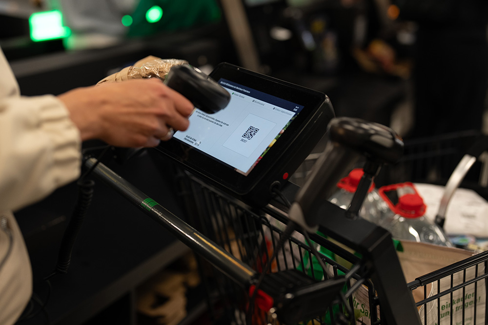Hand scannt mit einem Scanner an einem Einkaufswagen an einem Bildschirm, auf dem ein QR-Code angezeigt wird, im Supermarkt.