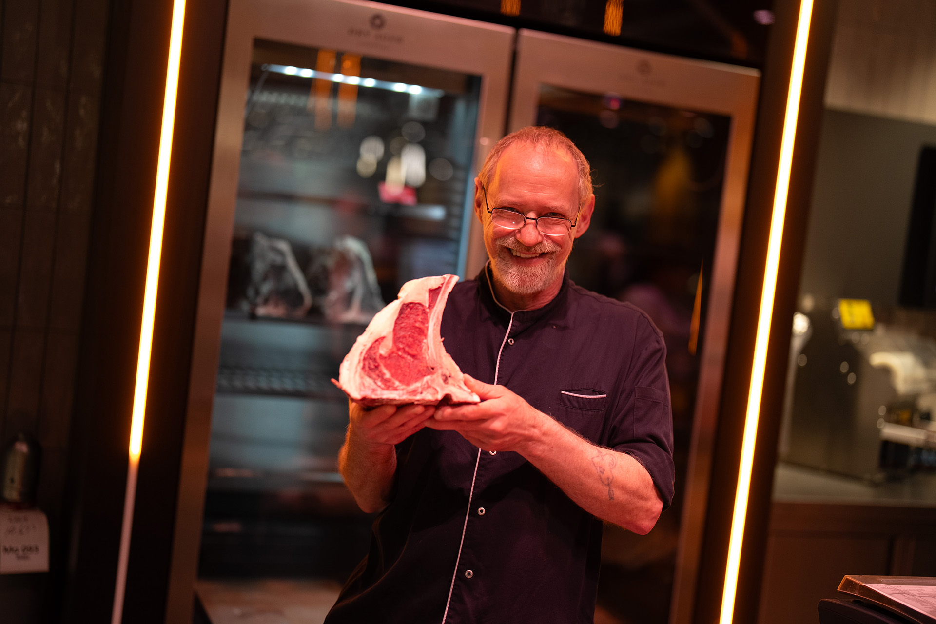 Ein älterer Mann mit Brille und Bart hält ein großes rohes Steak in beiden Händen und lächelt in einer Küche oder Metzgerei mit Kühlgeräten im Hintergrund.
