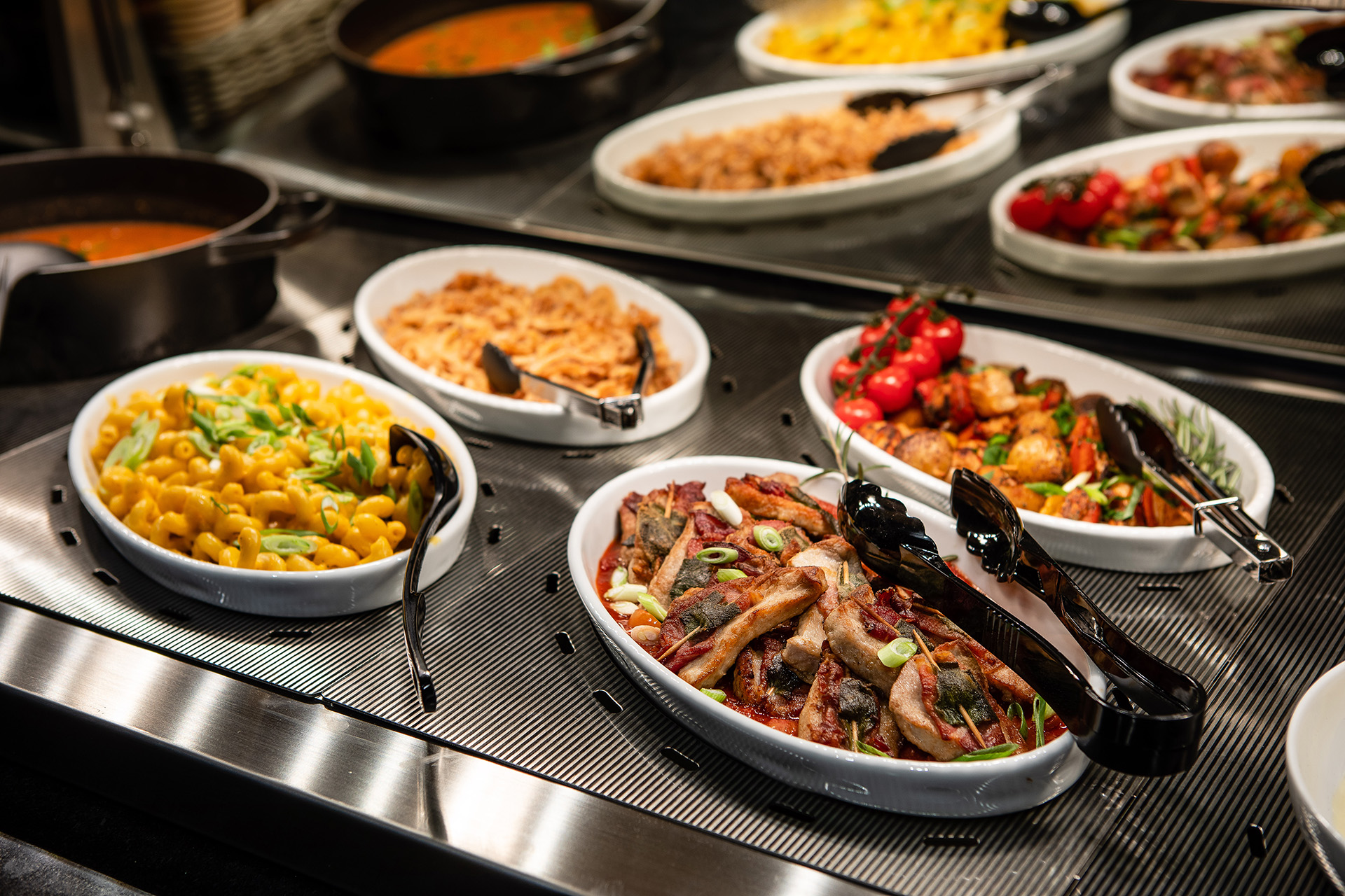 Buffet mit verschiedenen warmen Speisen in weißen Schalen, darunter Mac and Cheese mit Frühlingszwiebeln, gebratene Gemüse mit Tomaten und Fleischstücke mit Kräutern, alles auf einem beheizten Edelstahlbuffetständer.