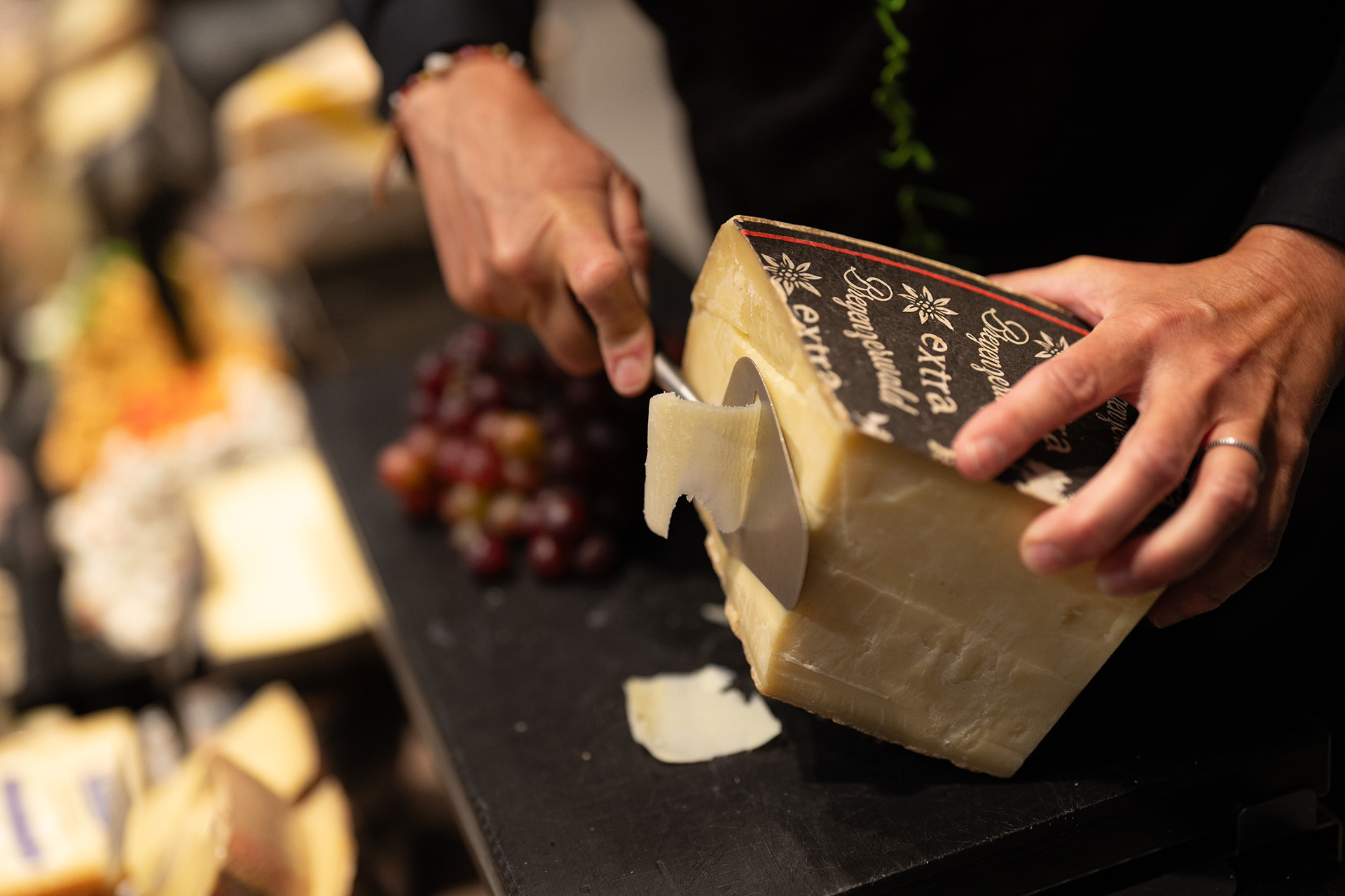 Person schneidet eine dünne Scheibe gereiften Käses von einem großen Klotz Käse mit einem speziellen Käseschaber, im Hintergrund unscharfe Trauben und weitere Käsestücke.