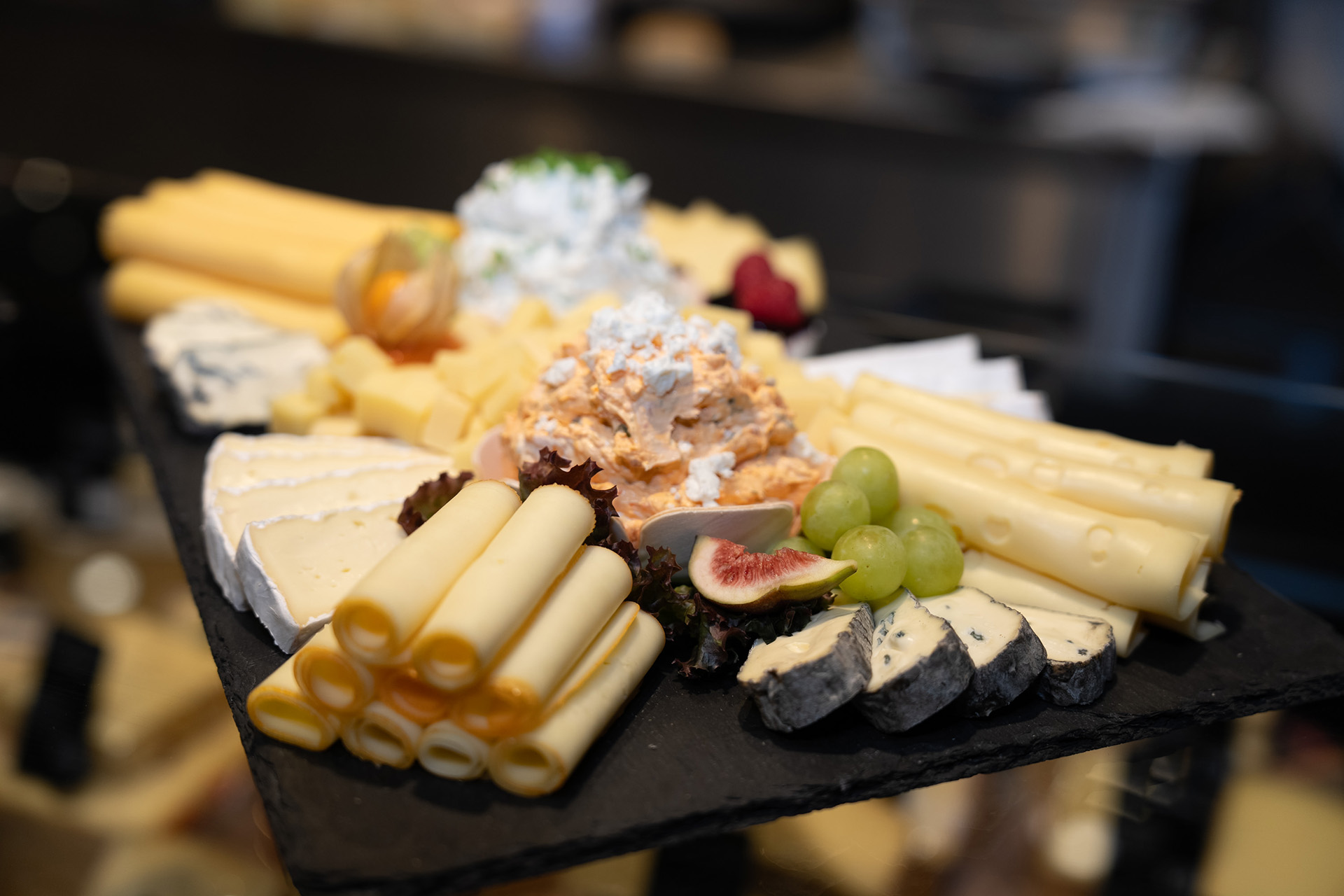 Verschiedene Käsesorten, darunter Käserollen und Blauschimmelkäse, auf einem schwarzen Servierbrett arrangiert mit Trauben, Feige und Cremeaufstrichen.