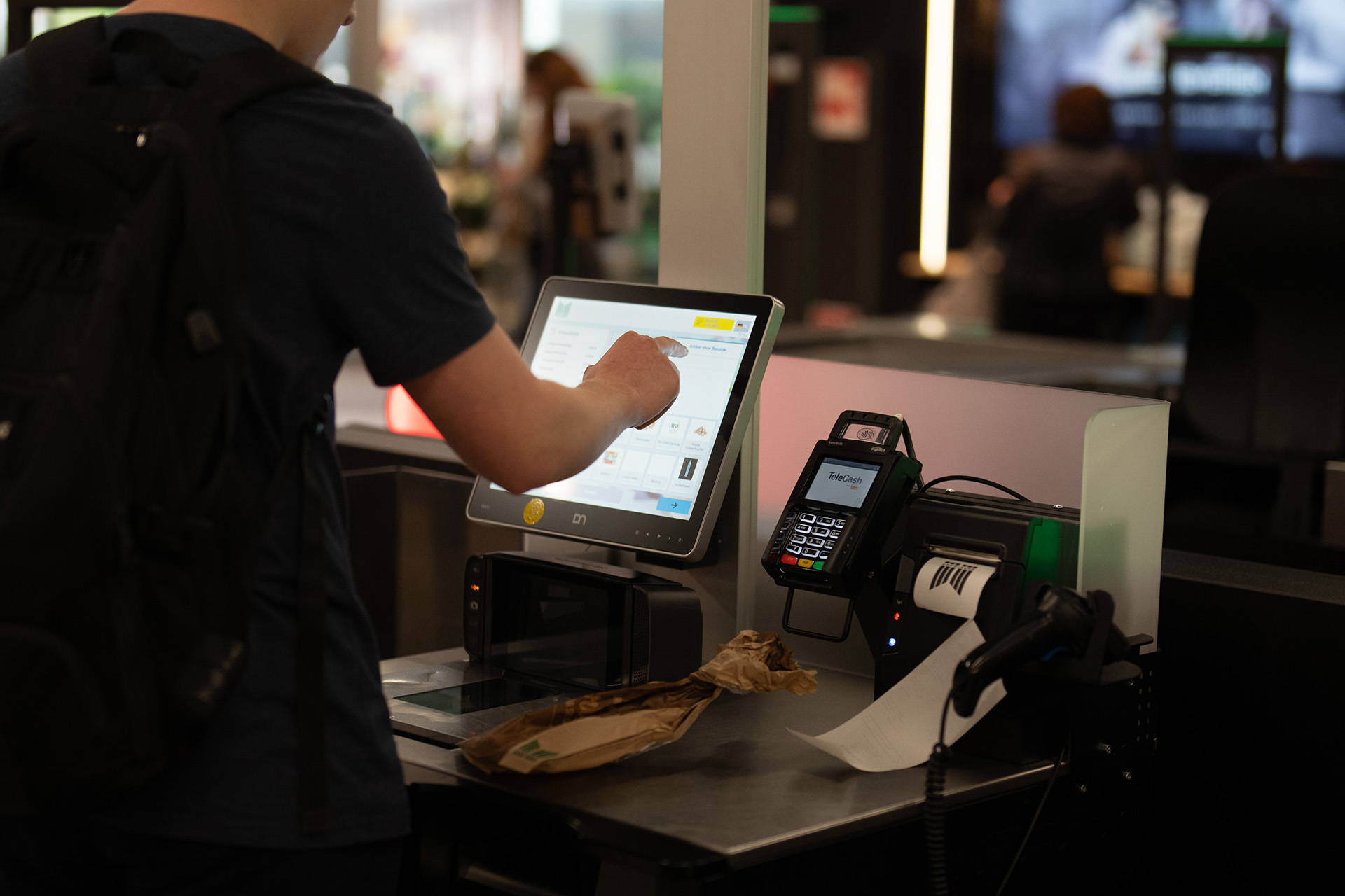 Person bedient einen Selbstbedienungskassen-Touchscreen mit schwarzem Rucksack, auf dem Kassenband liegt eine braune Papiertüte, daneben befinden sich ein Kartenlesegerät und ein Bonrollen-Drucker mit gedrucktem Beleg.