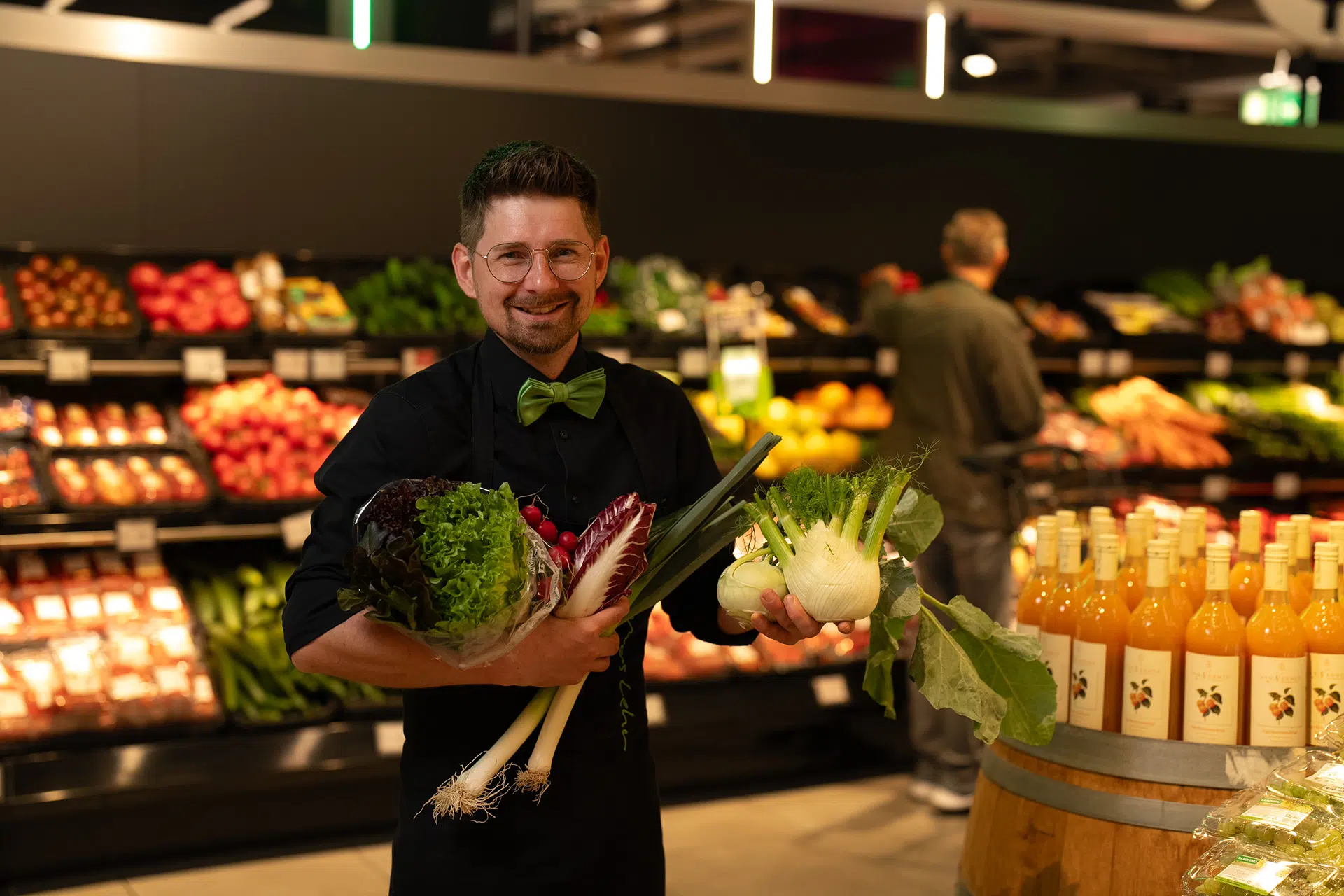 Mann mit Brille und grünem Fliegenhemd in einem Lebensmittelgeschäft hält frisches Gemüse wie Salat, Lauch und Fenchel in den Armen, im Hintergrund Regale mit Obst und Gemüse sowie ein weiterer Kunde.