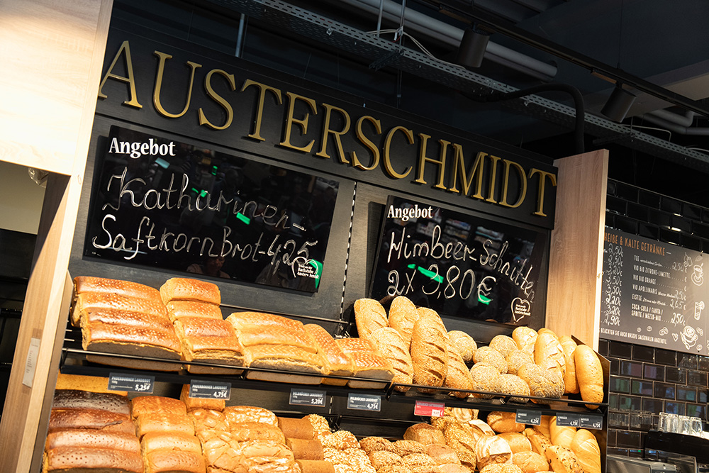 Bäckerei-Auslage mit verschiedenen Brotsorten unter der goldenen Aufschrift "AUSTERSCHMIDT" und zwei schwarzen Tafeln mit Angeboten für "Katharinen Saftkornbrot 4,25€" und "Himbeer-Schicht 2x 3,80€".