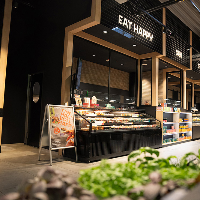 Modernes Sushi-Restaurant mit beleuchtetem Schriftzug "EAT HAPPY" und Auslagevitrine voller Sushi, im Vordergrund unscharfe grüne Pflanzen